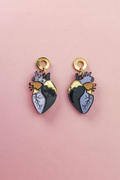 Two Anatomical Heart Earrings with gold, black, and blue details bring a hint of gothic flair, beautifully displayed on a pink background.