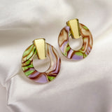 A pair of hypoallergenic Minimalist Circle Stud Earrings with gold rectangular tops and marbled acrylic glass circular bottoms in pink, green, and brown, displayed on white satin fabric.