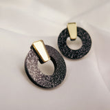 Minimalist Circle Stud Earrings crafted from hypoallergenic acrylic glass, these round black earrings feature silver speckles and gold geometric accents, displayed on a soft white fabric background.