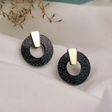 A pair of minimalist circle stud earrings in black acrylic glass with white speckles and gold triangular studs, displayed on white fabric with dried grasses in the background. Hypoallergenic for comfortable, everyday wear.