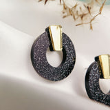 Close-up of minimalist circle stud earrings in black acrylic glass with silver glitter and a gold accent, displayed on a white fabric background with blurred dried plant details above. Hypoallergenic design ensures comfortable wear.
