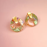 A pair of minimalist circle stud earrings crafted from acrylic glass in marbled pastel green, pink, and purple, with gold hypoallergenic rectangular studs, displayed on a soft pink background.