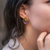 A close-up of a woman wearing minimalist circle stud earrings crafted from acrylic glass, featuring abstract multicolored patterns and a gold rectangular accent. She has dark hair and is touching her chin with her hand.