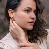 A woman with wavy brown hair wearing a beige top touches her Minimalist Circle Stud Earrings with one hand and rests her other hand on her collarbone. She looks slightly to the side with a calm expression. The earrings are hypoallergenic.