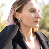 A woman with light brown hair touches her head while looking into the distance. She wears a black coat, a mauve top, and Minimalist Circle Stud Earrings that are hypoallergenic. The background is outdoors and softly blurred by sunlight.