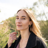 A woman with long, light brown hair stands outdoors, wearing a dark coat, mauve top, and minimalist circle stud earrings. She lightly touches her hair and looks calmly at the camera against green foliage and a bright, sunlit sky.