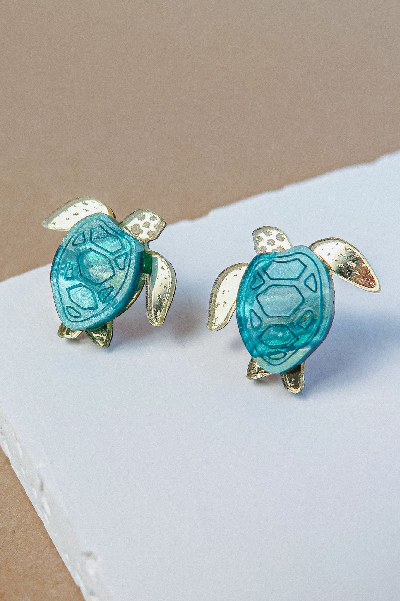 Sea Turtle Studs shaped like turtles with turquoise blue shells and gold-toned legs, heads, and fins bring a summery vibe, displayed on a white and beige background.