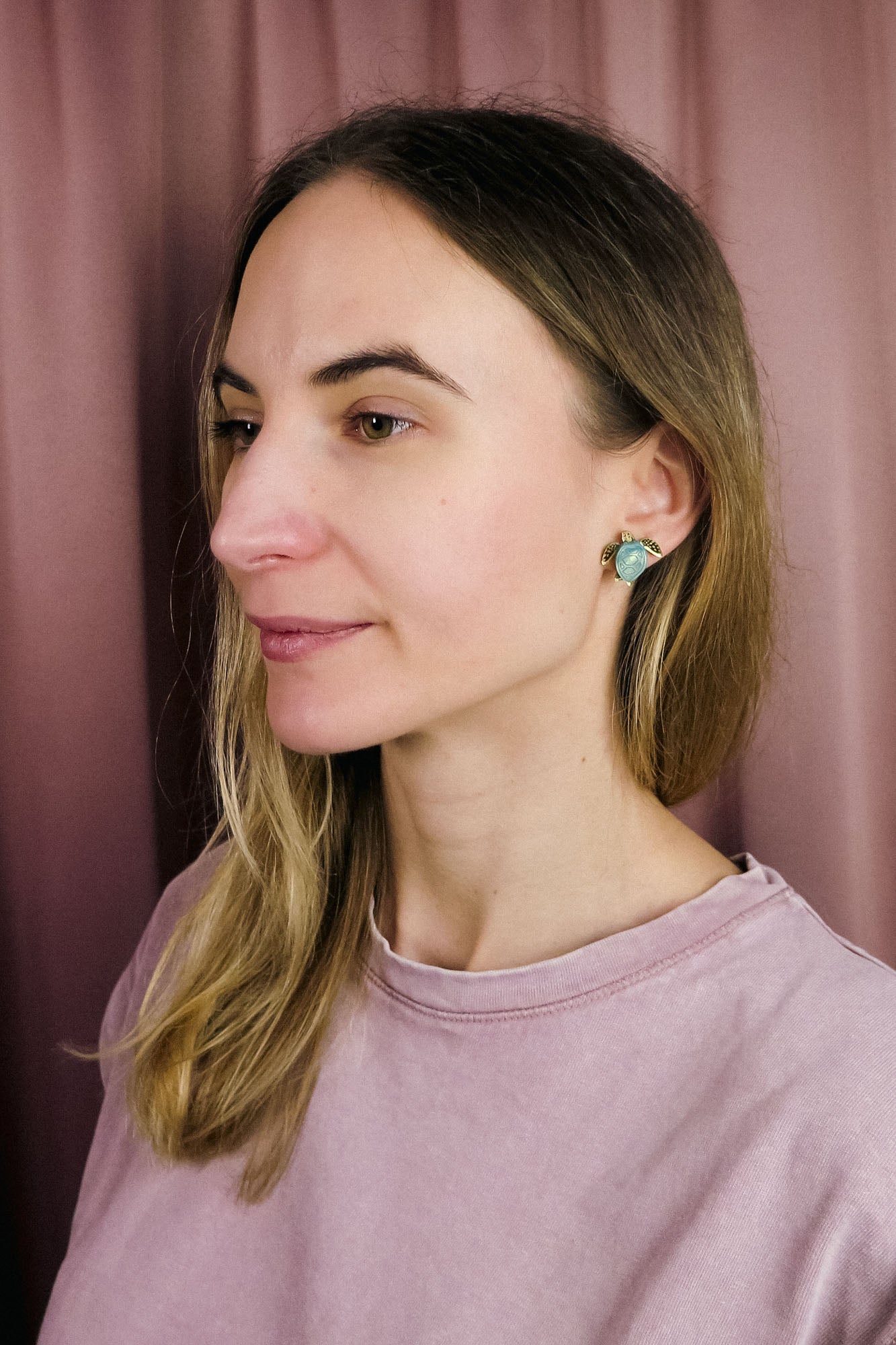 A woman with straight, light brown hair and light skin wears a light pink top and turquoise Sea Turtle Studs, standing in front of a mauve curtain, gazing slightly to the left for a summery vibe.