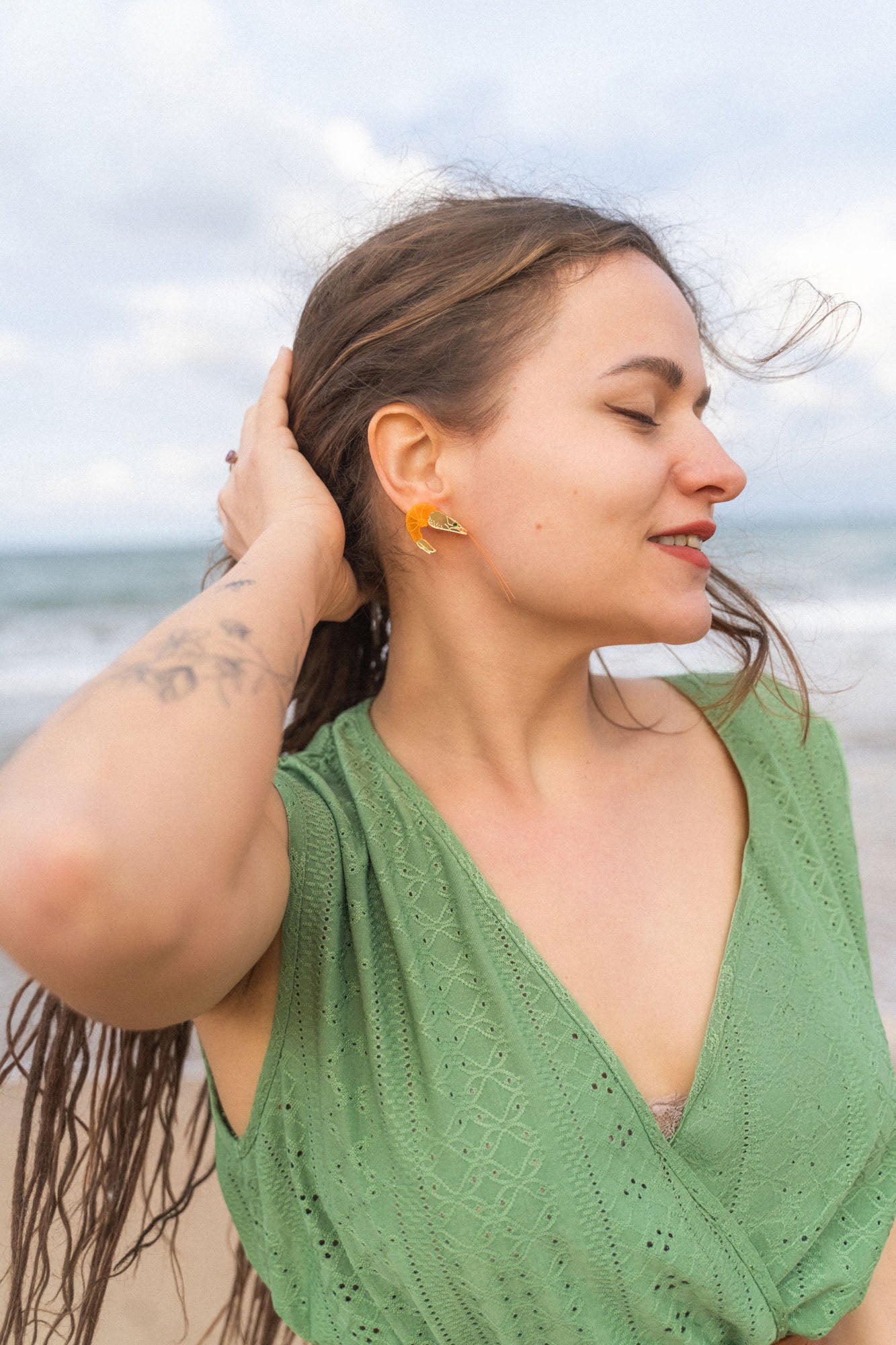 A woman with long brown hair in a green dress stands on a beach, eyes closed and smiling slightly, with one hand touching her hair. She wears neon orange Shrimp Stud earrings and has a tattoo on her arm. The ocean is in the background.