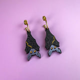 A pair of hand-painted bat-shaped earrings with black, glittery wings and gold accents, hanging from gold hoops, displayed on a pastel purple background. These allergy-free earrings combine unique design with comfortable wear.