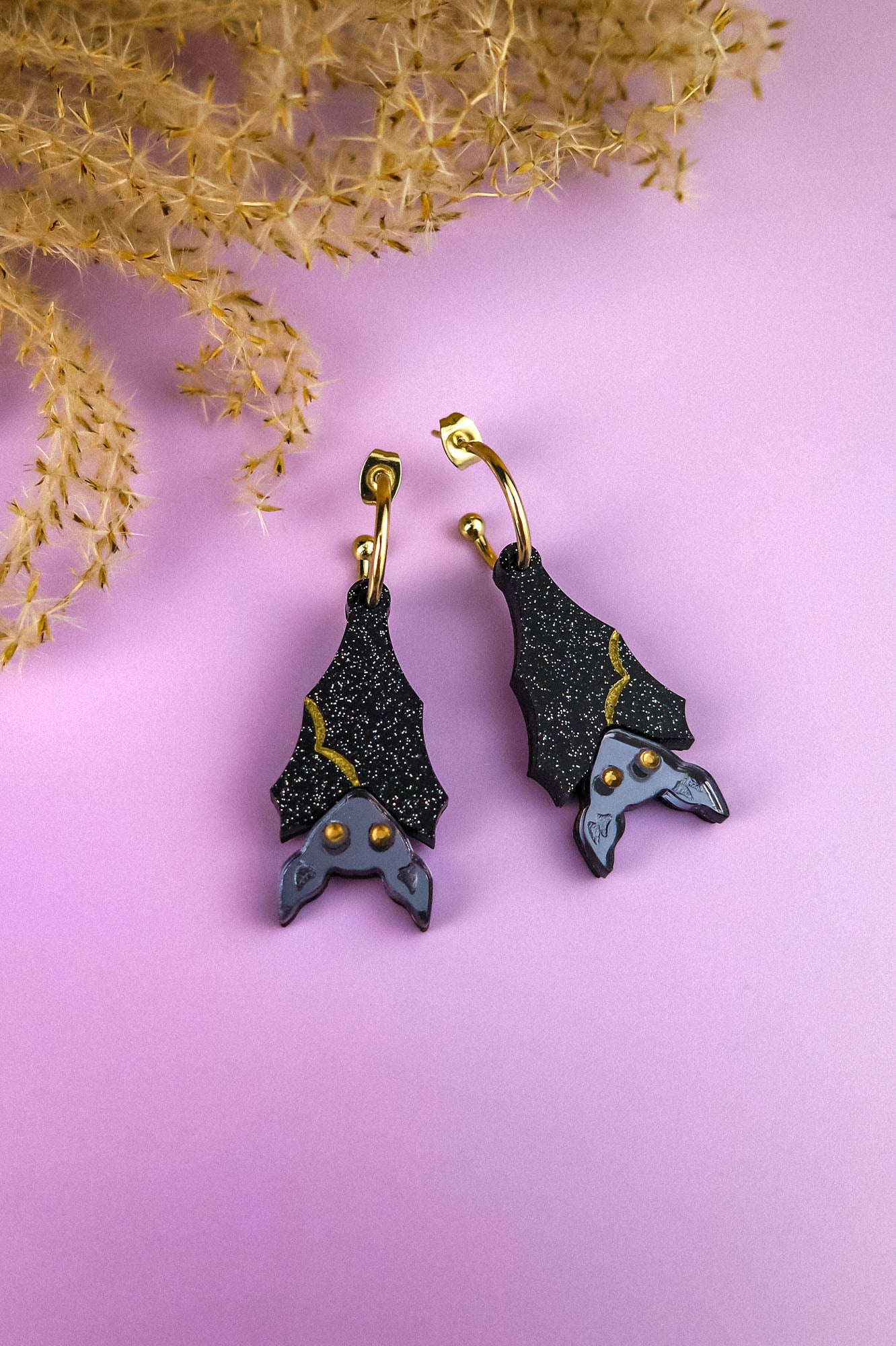 A pair of hand-painted, black bat-shaped earrings with gold accents and gray faces, displayed on a pastel purple surface with dried beige foliage in the corner.