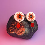 A pair of hand-painted earrings designed like bloodshot eyeballs with red teardrop-shaped pendants hang from a dark, textured rock against a purple background.