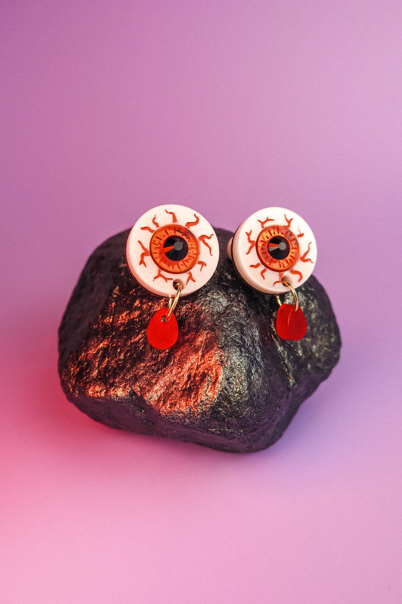 A pair of hand-painted earrings designed like bloodshot eyeballs with red teardrop-shaped pendants hang from a dark, textured rock against a purple background.