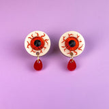 Two hand-painted earrings designed as white eyeballs with red veins and black pupils, each featuring a dangling red teardrop, are displayed on a light purple background. These allergy-free earrings add a quirky, artistic touch to any look.