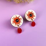 A pair of hand-painted earrings shaped like bloodshot eyeballs with red veins and dangling red teardrops, displayed on a lavender surface with dried flowers in the background.