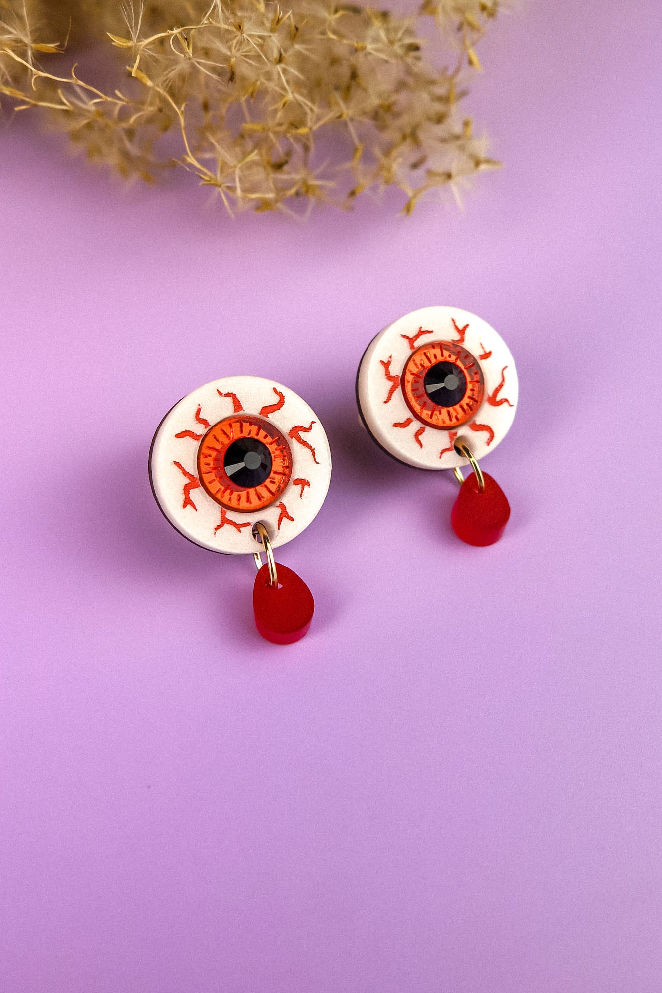 A pair of hand-painted earrings shaped like bloodshot eyeballs with red veins and dangling red teardrops, displayed on a lavender surface with dried flowers in the background.