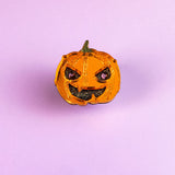 A small, shiny, orange Spooky Pumpkin Pin with a green stem and a mischievous expression sits on a plain light purple background, adding handcrafted Halloween charm to any décor.