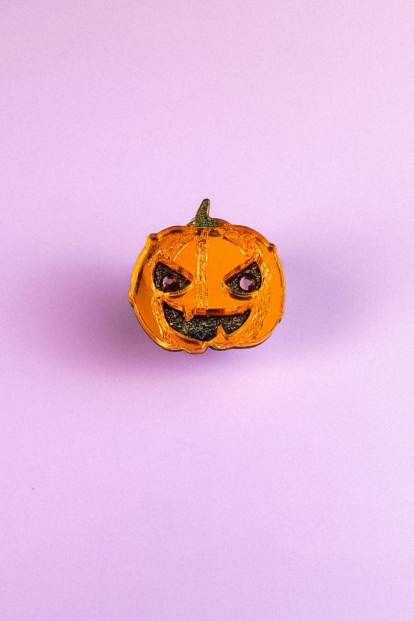 A small, shiny, orange Spooky Pumpkin Pin with a green stem and a mischievous expression sits on a plain light purple background, adding handcrafted Halloween charm to any décor.