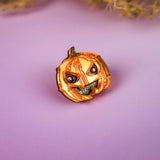 A small, handcrafted Spooky Pumpkin Pin with a mischievous face sits on a purple surface, perfect for adding a touch of Halloween charm amid blurred dried plant decorations in the background.