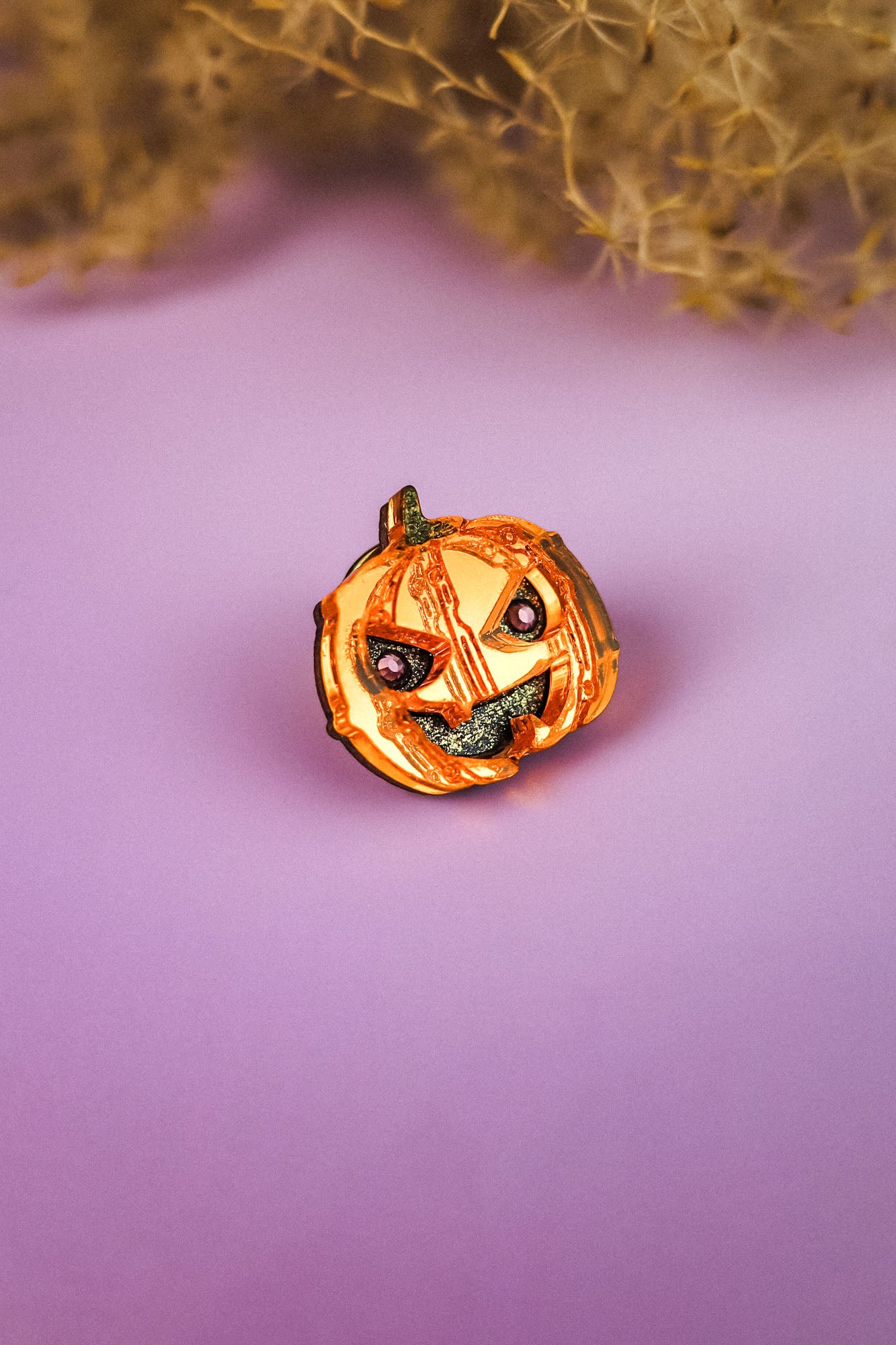 A small, handcrafted Spooky Pumpkin Pin with a mischievous face sits on a purple surface, perfect for adding a touch of Halloween charm amid blurred dried plant decorations in the background.