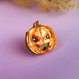 A small, handcrafted Spooky Pumpkin Pin with a mischievous face is displayed on a smooth, purple surface with blurred beige foliage in the background—perfect for adding Halloween flair to your outfit.