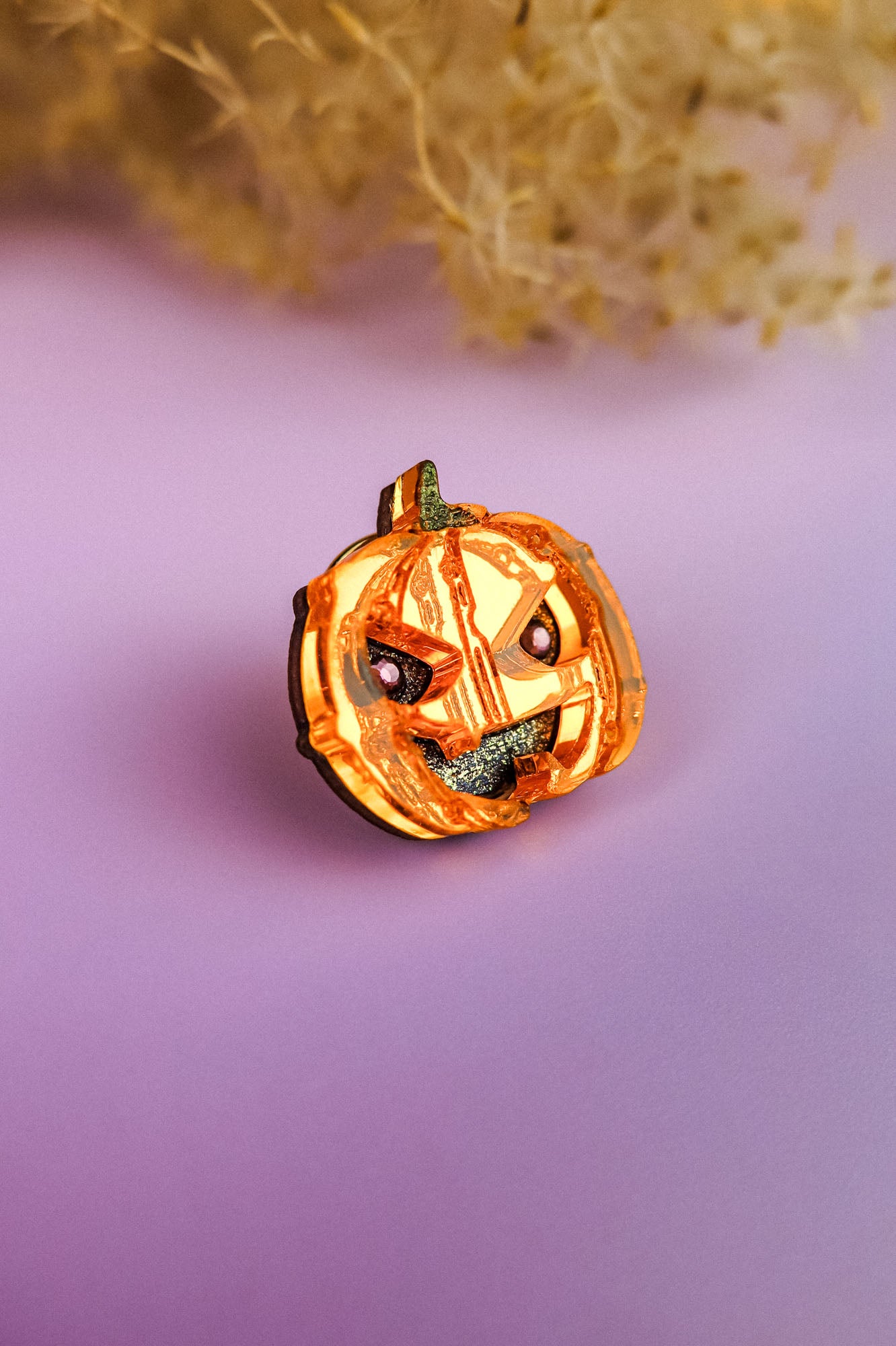 A small, handcrafted Spooky Pumpkin Pin with a mischievous face is displayed on a smooth, purple surface with blurred beige foliage in the background—perfect for adding Halloween flair to your outfit.