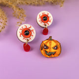 Two handcrafted white earrings shaped like bloodshot eyeballs with red teardrop charms and a spooky pumpkin pin complete this Halloween look, all arranged on a purple surface with dried foliage in the background.