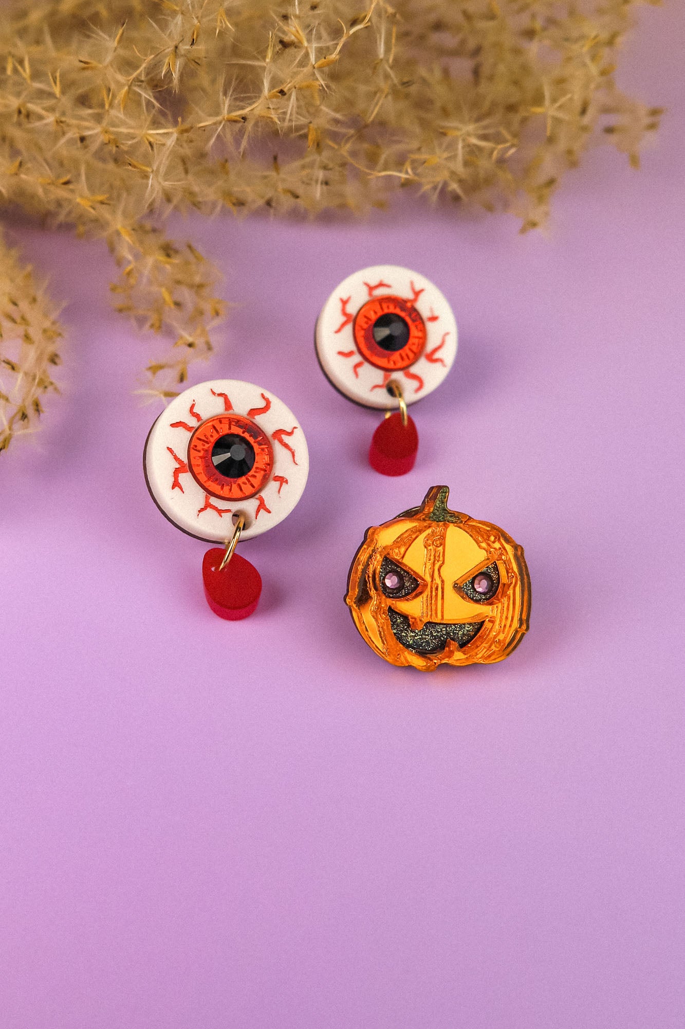 Two handcrafted white earrings shaped like bloodshot eyeballs with red teardrop charms and a spooky pumpkin pin complete this Halloween look, all arranged on a purple surface with dried foliage in the background.