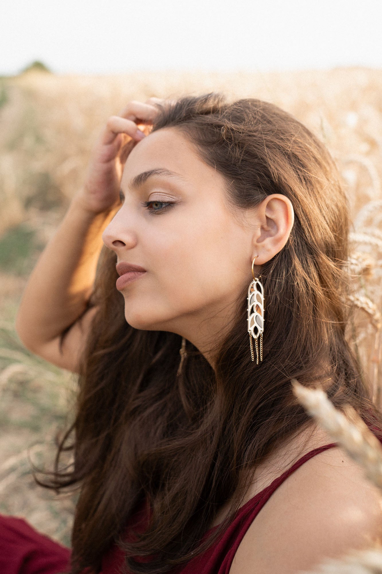 A young woman with long brown hair sits in a wheat field, wearing a burgundy dress and Wheat Statement Earrings. She gazes thoughtfully to the side, lightly touching her hair, her standout style enhanced by the bold design of her earrings.