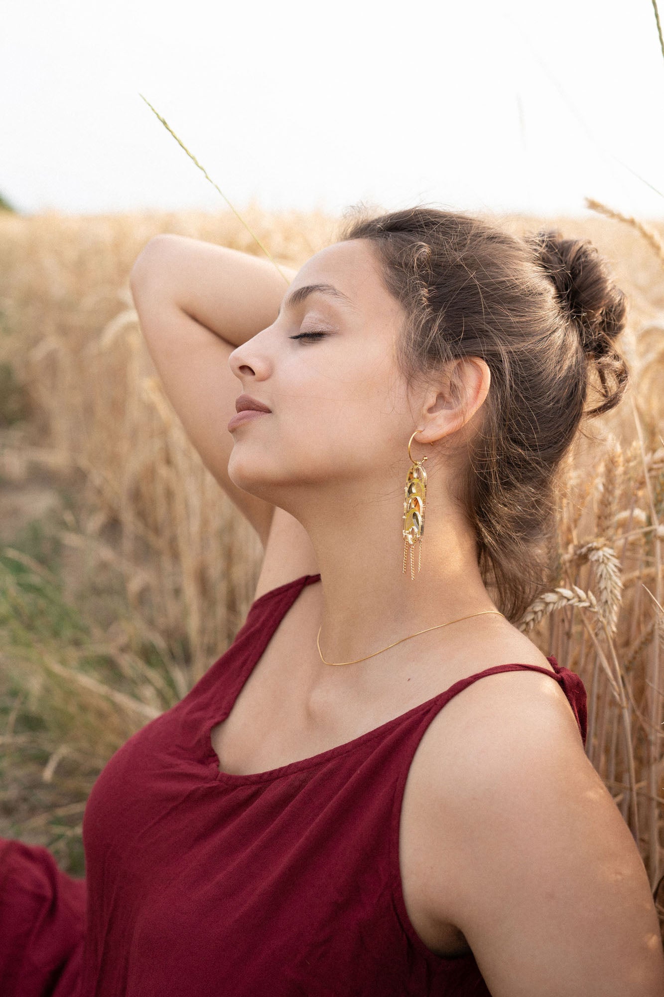 A young woman in a burgundy dress sits in a wheat field with her eyes closed, one arm raised behind her head. She wears Wheat Statement Earrings and a delicate necklace, appearing relaxed and peaceful in the natural setting with standout style.