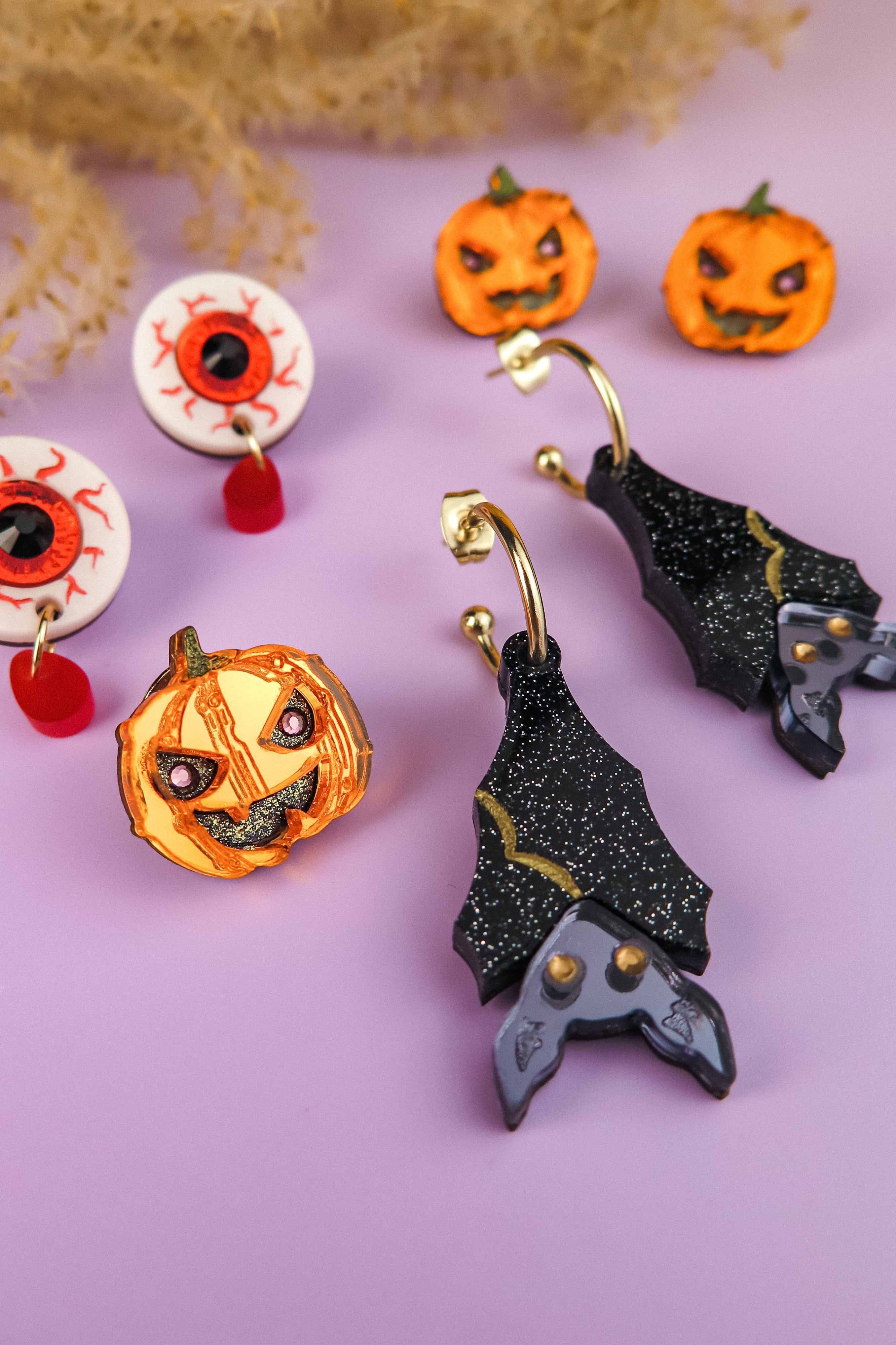Assorted Halloween-themed earrings and pins on a purple surface, featuring jack-o-lanterns, eyeballs, and black bat wings with gold and silver glitter accents. Dried foliage decor in the background.