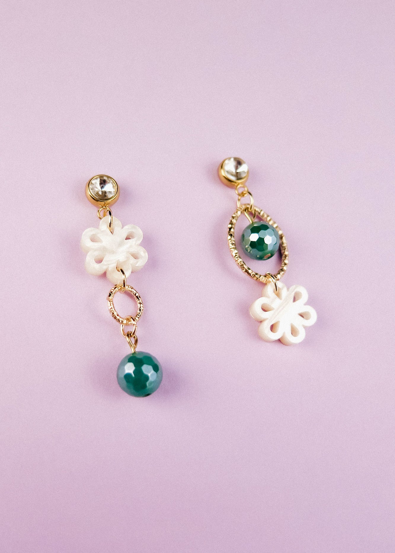 A pair of mismatched earrings on a light purple background; both feature white floral designs, gold accents, and green beads—blending modern charm with timeless elegance in their circular and oval shapes.