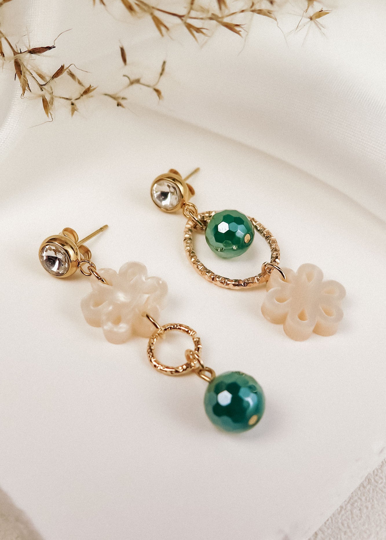 A pair of mismatched gold earrings with green beads and cream flower-shaped charms exudes timeless elegance, displayed on a white surface with dried grass in the background.