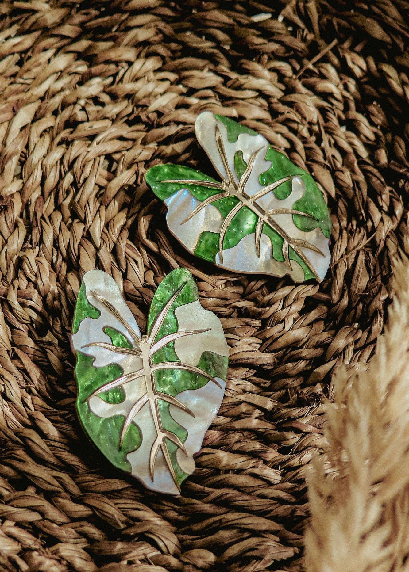 Two decorative brooches shaped like green monstera leaves with white accents rest on a woven natural fiber surface, resembling hand-crafted jewelry. Beige pampas grass adds a soft touch in the lower right corner.