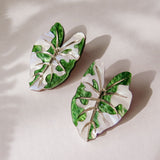 Two Alocasia Earrings shaped like monstera leaves with green and white enamel detailing are placed on a light surface, casting soft shadows—perfect for those who love lightweight jewelry.