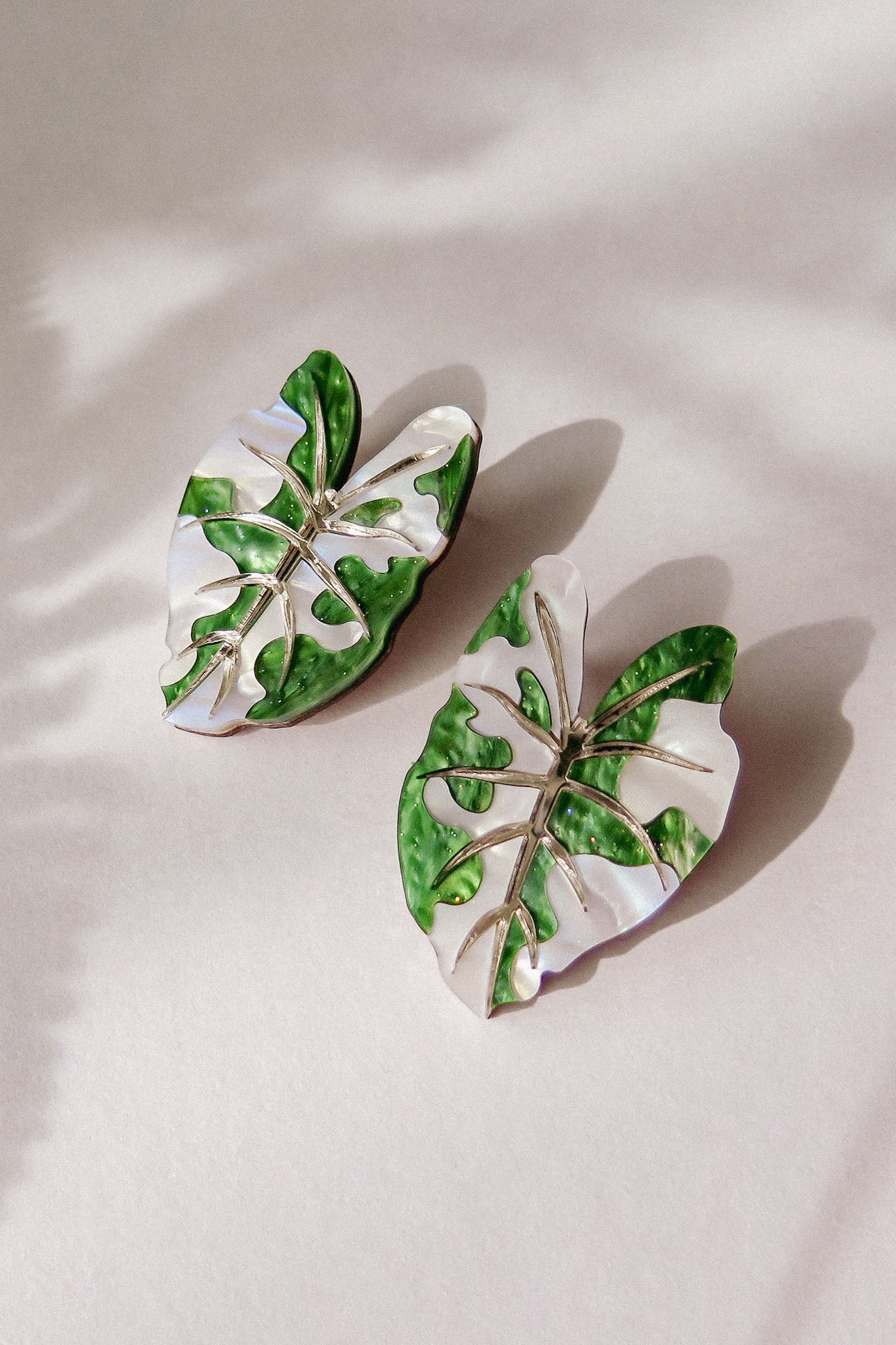 Two Alocasia Earrings shaped like monstera leaves with green and white enamel detailing are placed on a light surface, casting soft shadows—perfect for those who love lightweight jewelry.