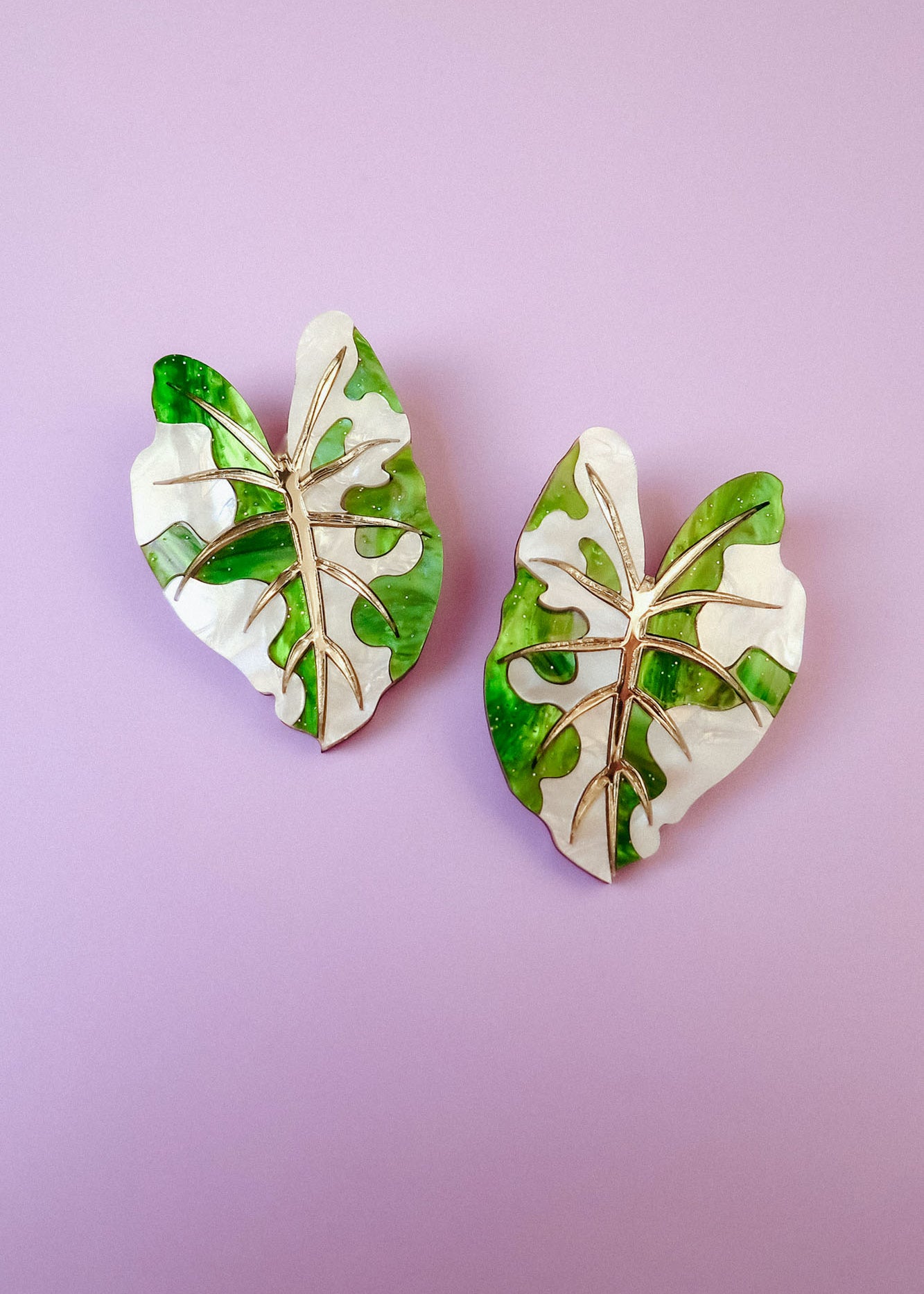 Two decorative pins shaped like monstera leaves, inspired by hand-crafted jewelry, feature a mix of green and white colors with gold vein detailing, and are placed on a light purple background.