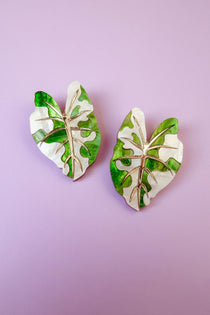 Two decorative pins shaped like monstera leaves, inspired by hand-crafted jewelry, feature a mix of green and white colors with gold vein detailing, and are placed on a light purple background.