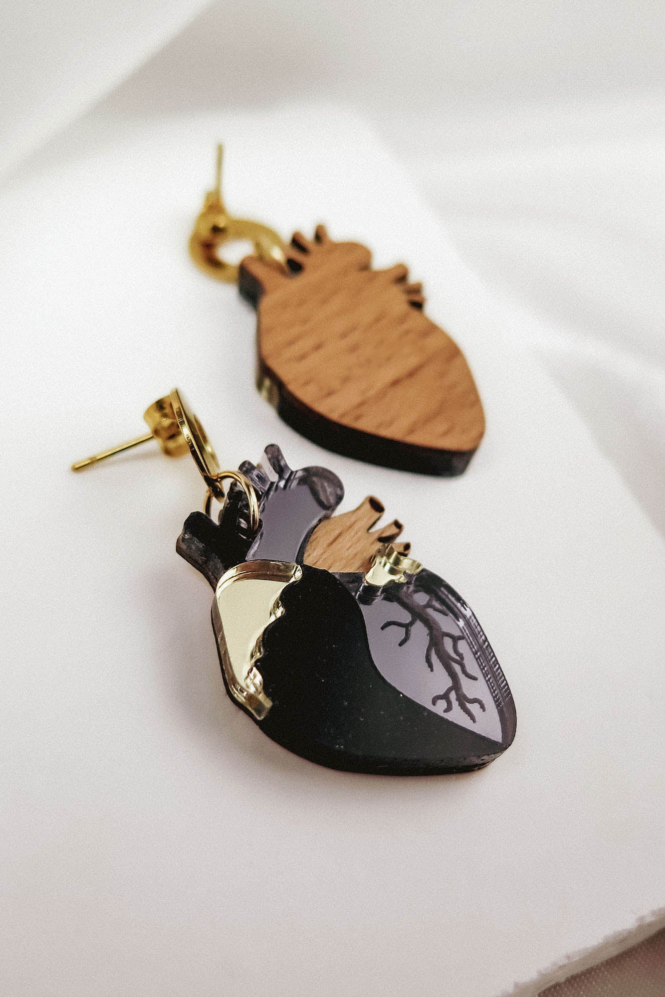 A close-up of two Anatomical Heart Earrings, handcrafted from wood and black acrylic with gold hooks, showcases unique handcrafted accessories and a hint of gothic flair on a white surface.