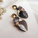 Two anatomical heart earrings with black, silver, and gold accents add a touch of gothic flair, lying on white fabric beside delicate dried flowers—unique handcrafted accessories for those who love bold style.