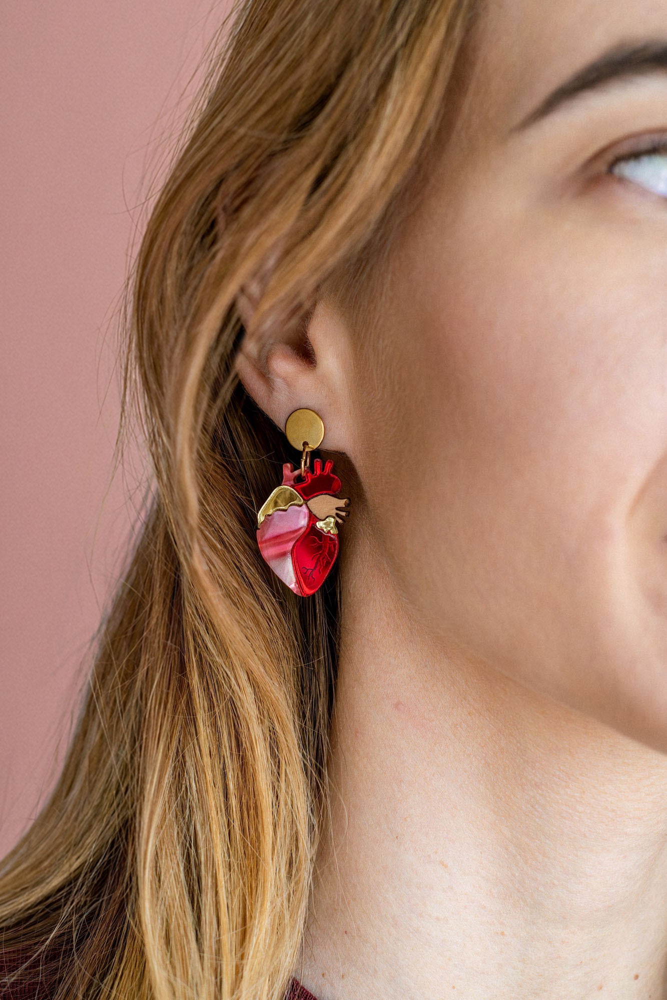 Close-up of a woman’s ear wearing unique style Anatomical Heart Earrings in red and pink tones with gold accents; her light brown hair is tucked behind her ear against a pink background, highlighting these striking acrylic earrings.