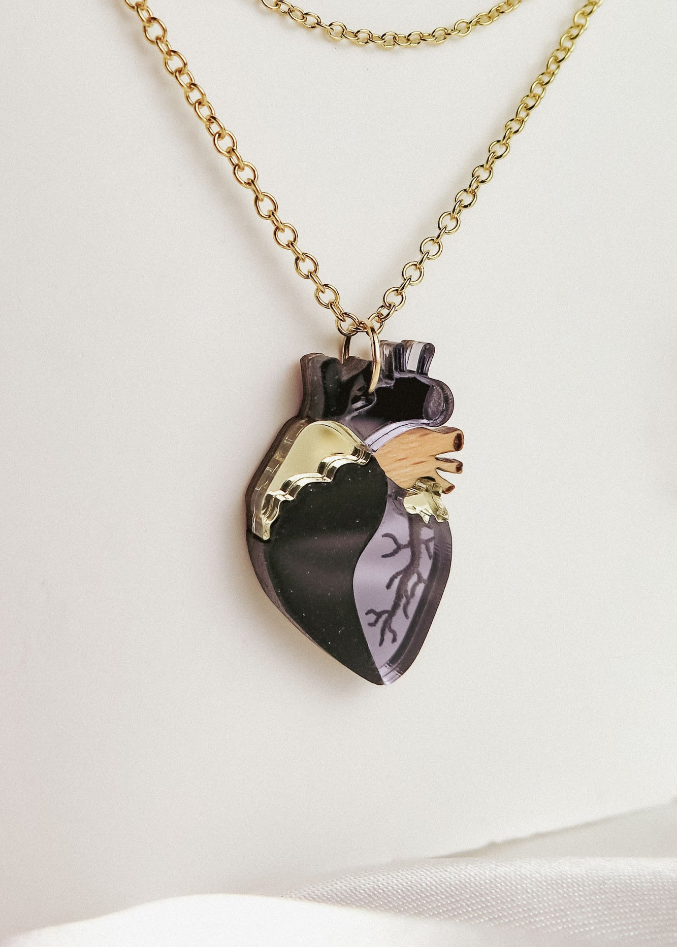 A gold chain necklace with a pendant shaped like an anatomical heart, featuring black, gold, and wood-toned sections etched with vein-like details. This handcrafted jewelry piece rests against a white background.