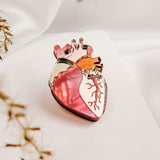 A unique handcrafted glass object shaped like an anatomical heart, featuring shades of pink and gold, sits on a soft white surface with blurred dried plant stems in the foreground.