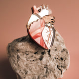 A unique, handcrafted Anatomical Heart Pin in metallic pink rests on a rough, light-colored rock against a soft, warm-toned background.