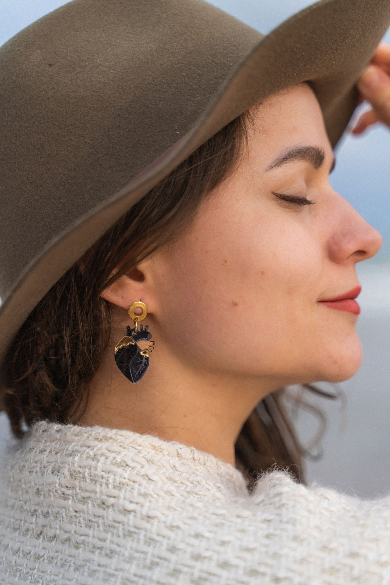 A woman in a brown hat and cream sweater smiles with her eyes closed, showing off handcrafted accessories—a black and gold leafy earring. The background is softly blurred.