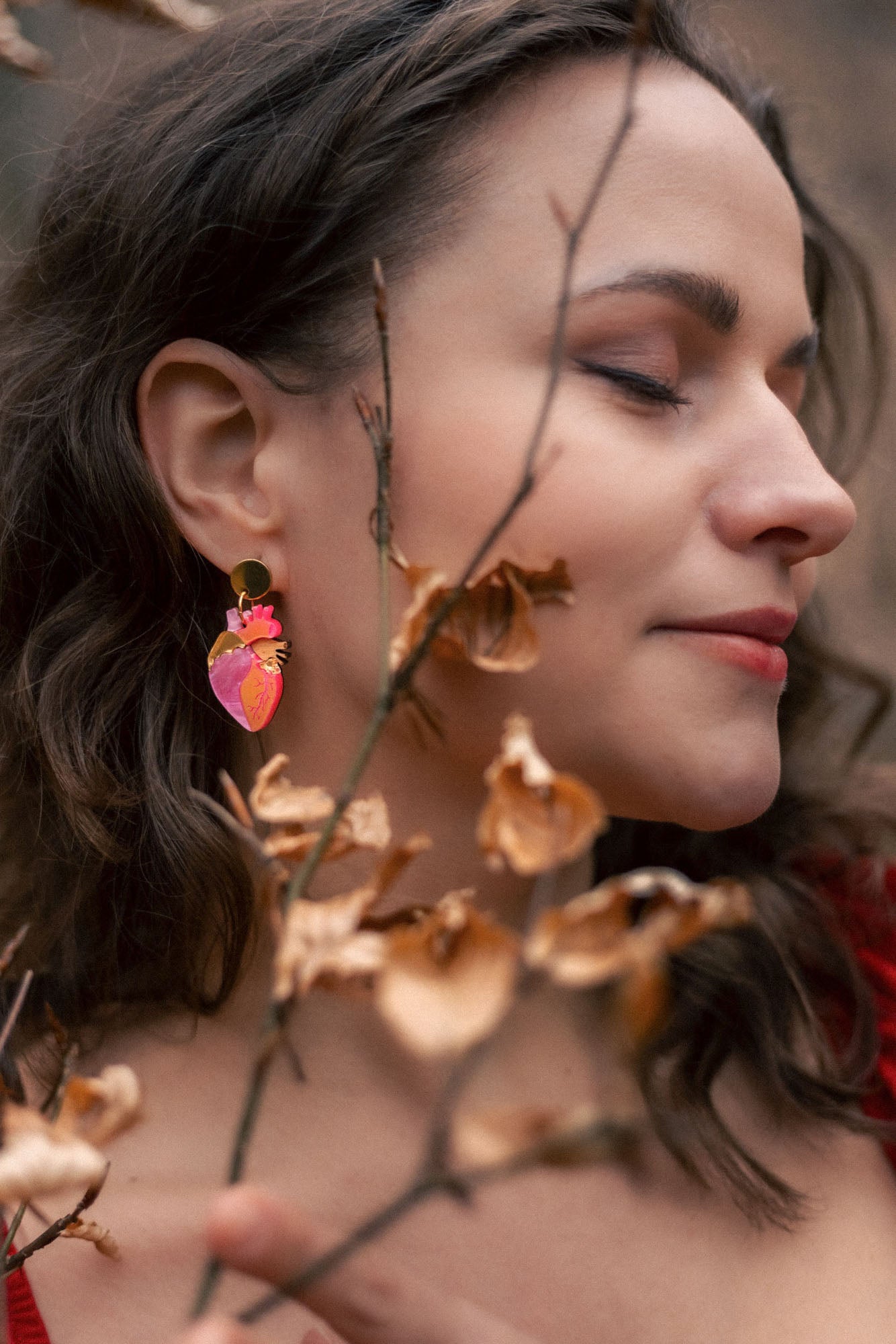 A woman with wavy brown hair and closed eyes stands peacefully among dry brown branches, wearing bold style pink and gold earrings and a red top.