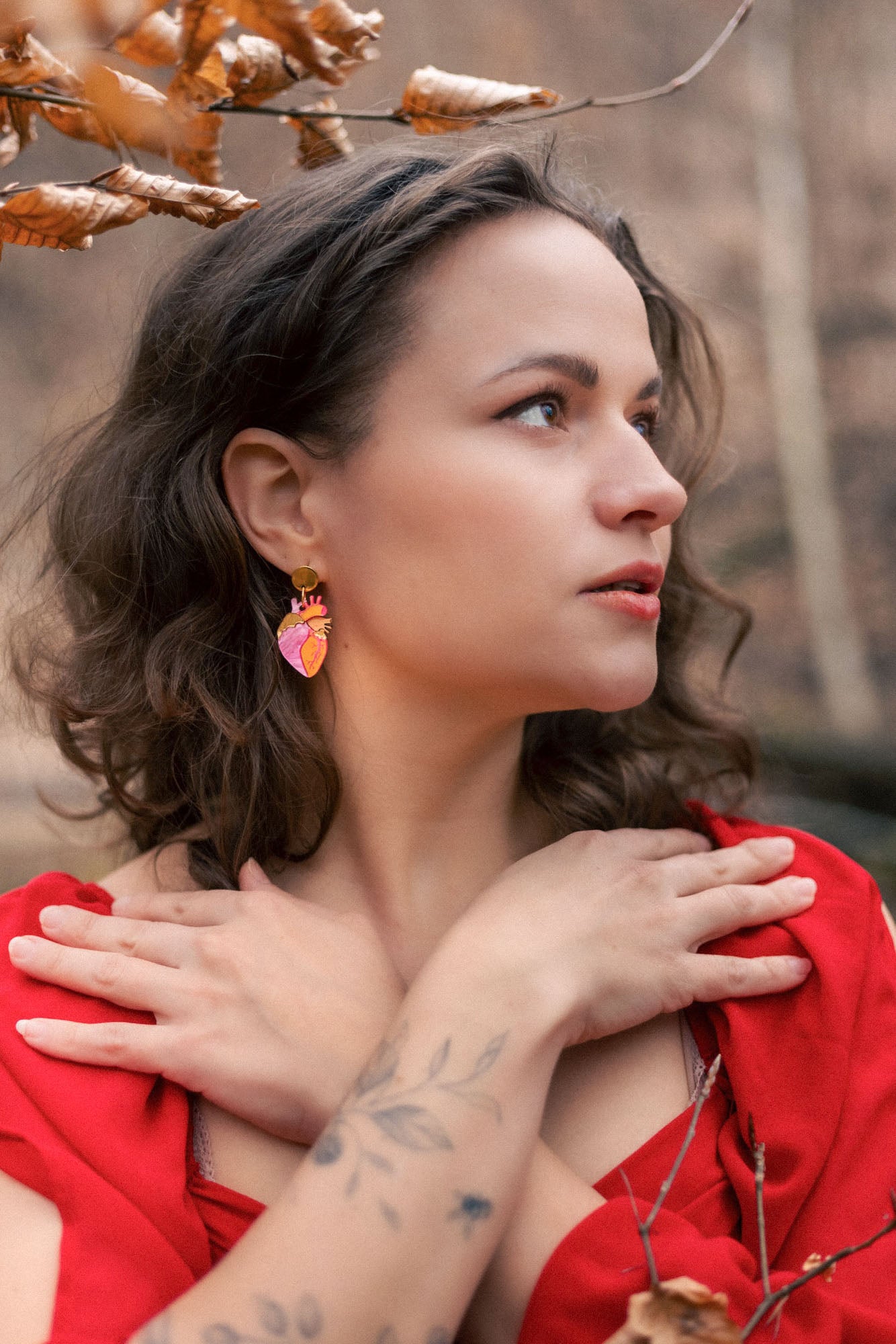 A woman with wavy brown hair and a tattooed forearm crosses her hands over her chest. She wears a red dress, bold style pink statement earrings, and stands outdoors among brown autumn leaves, looking to the side.
