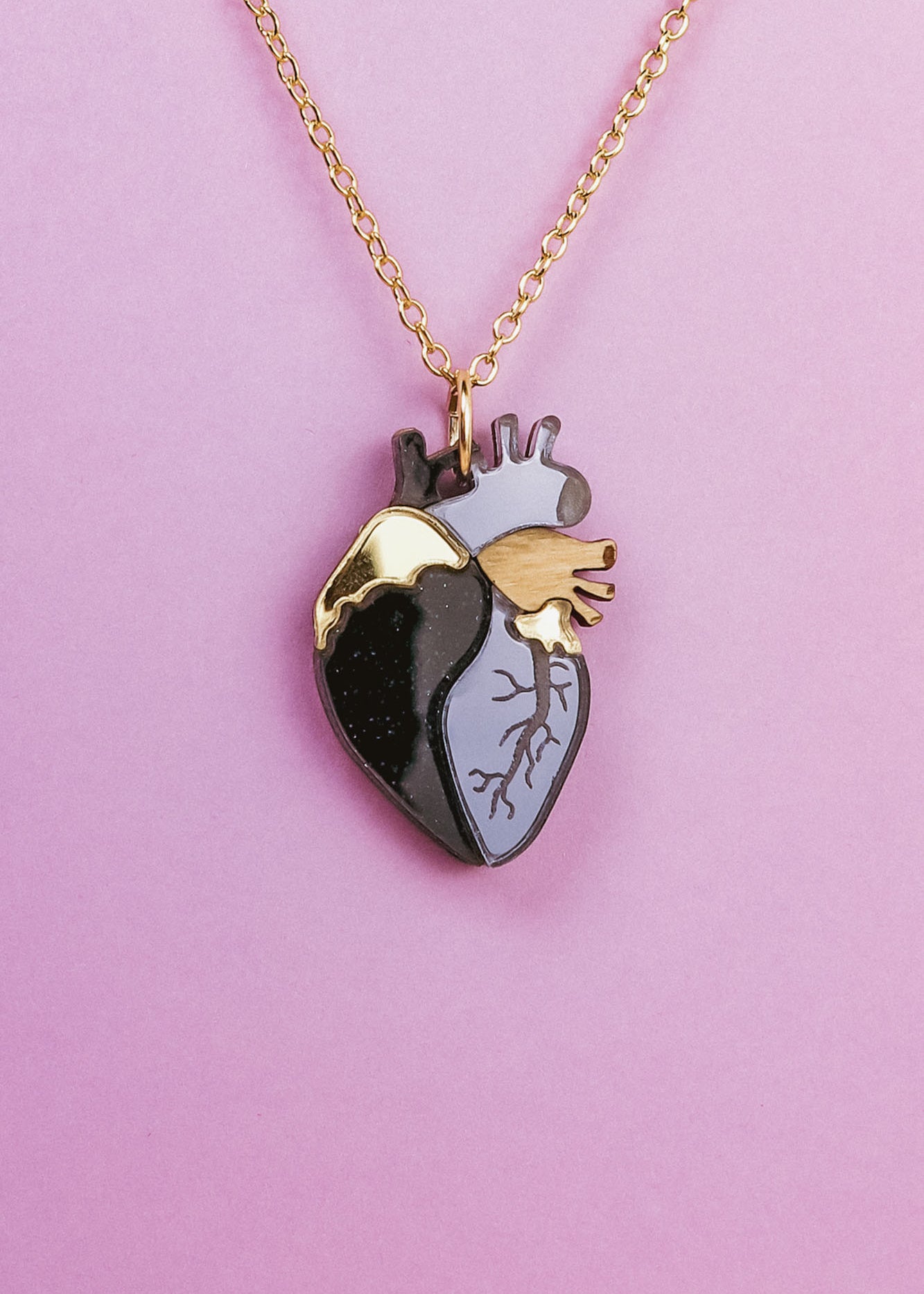 A gold chain anatomical heart necklace with a pendant featuring black, gray, and gold sections is displayed against a pink background—a truly unique jewelry piece.