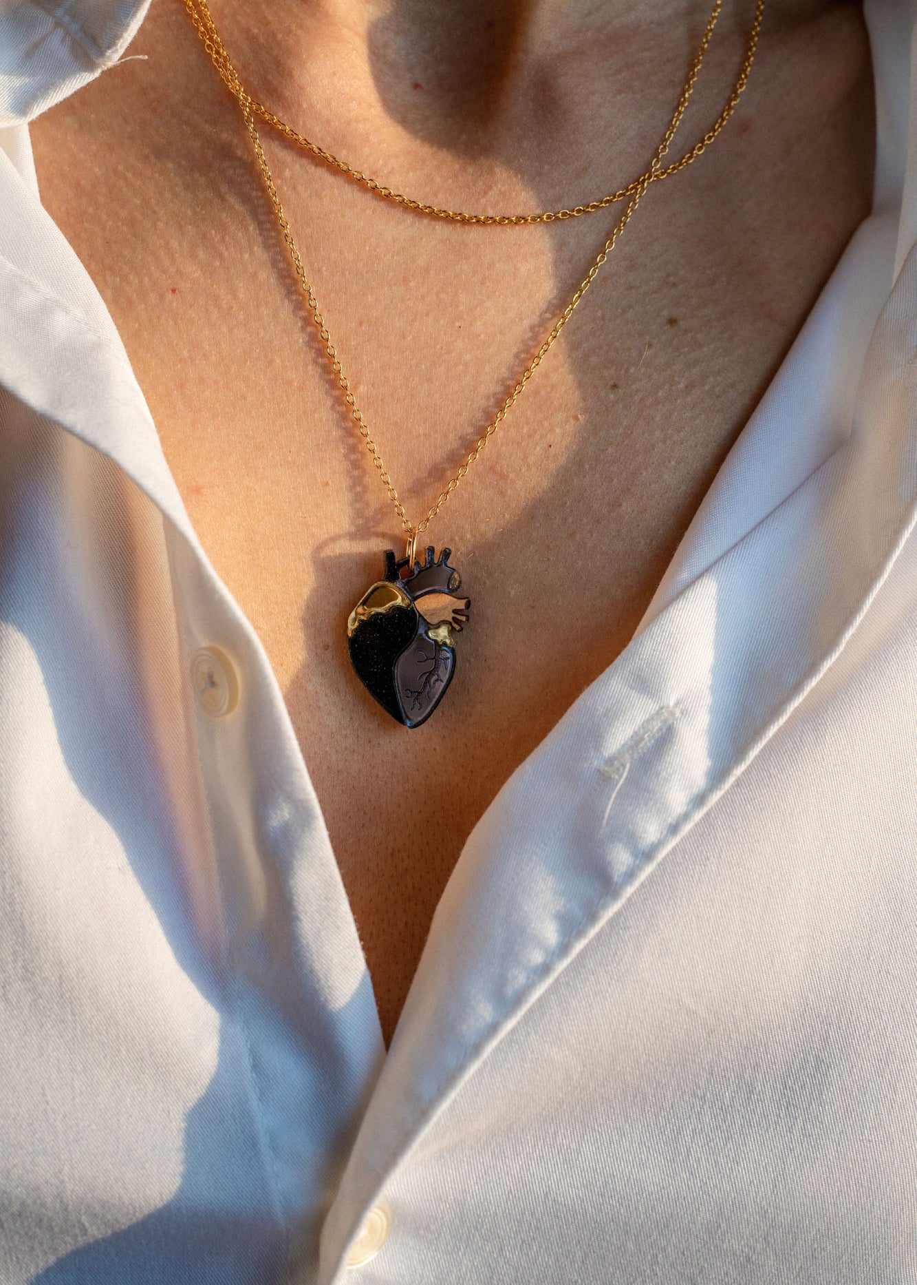 A person wearing a white button-up shirt with two layered gold necklaces, including a unique anatomical heart necklace. The scene is lit by warm sunlight.