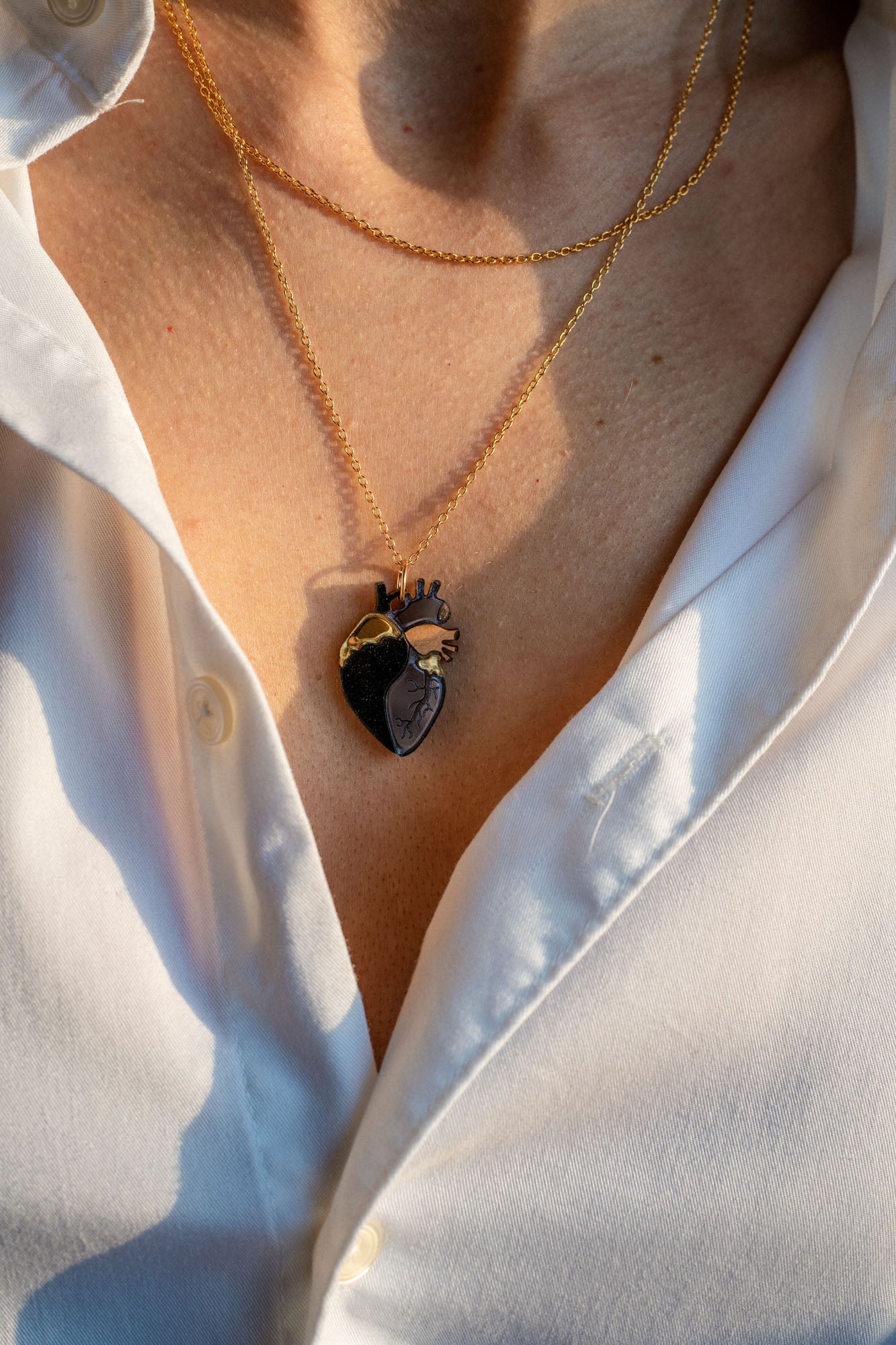 A person wearing a white button-up shirt with two layered gold necklaces, including a unique anatomical heart necklace. The scene is lit by warm sunlight.